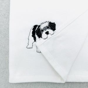 Könnte beinhalten: Weißer Stoff mit einem gestickten Hundemotiv in Schwarz und Weiß. Der Hund ist eine kleine Rasse mit flauschigem Aussehen. Der Stoff hat ein subtiles Fischgrätenmuster. Der Hund ist mit schwarzem Faden gestickt.