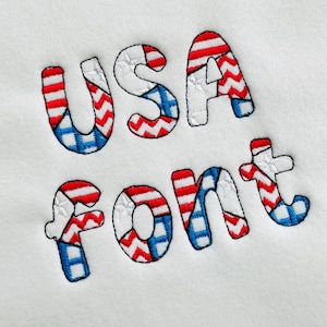 Könnte beinhalten: Gestickter Text "USA font" auf weißem Hintergrund. Die Buchstaben sind rot, weiß und blau, mit Mustern, die an die amerikanische Flagge erinnern. Die Schriftart ist ein verspieltes, gesticktes Design.