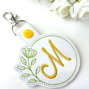May include: White leather keychain with a yellow button and a green embroidered floral design. The keychain features a white circle with a green outline and a gold embroidered letter "M" in the center.