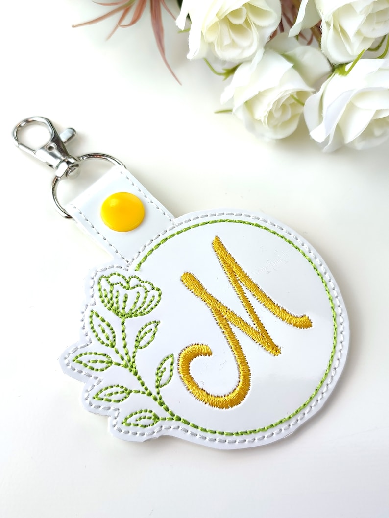 May include: White leather keychain with a yellow button and a green embroidered floral design. The keychain features a white circle with a green outline and a gold embroidered letter "M" in the center.