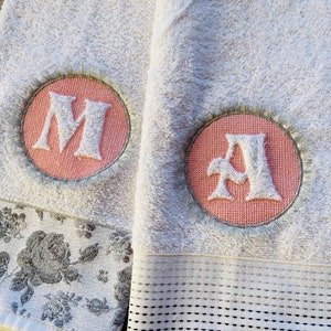 May include: Two white towels with embroidered pink circles containing the letters "M" and "A" in white. The towels have a decorative border with a floral pattern.