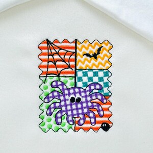 May include: Embroidered Halloween design featuring a purple spider with a checkered body, a spiderweb, and a bat. The design is on a white background and is composed of four colorful patchwork squares with orange, green, and yellow accents.