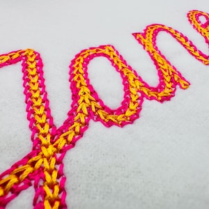 Könnte beinhalten: Nahaufnahme des Namens "Jane", gestickt in einem kursiven Stil. Die Buchstaben sind ein leuchtendes Pink, umrandet von einem goldgelben Faden, was einen auffälligen Kontrast zum weißen Stoffhintergrund erzeugt.