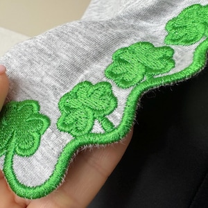 Puede incluir: Tela gris con borde festoneado y tréboles bordados en verde.