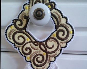 Puede incluir: Una toalla blanca con un diseño bordado en marrón y amarillo. El diseño presenta remolinos y puntos. La toalla está colgada en un pomo de puerta de armario blanco.