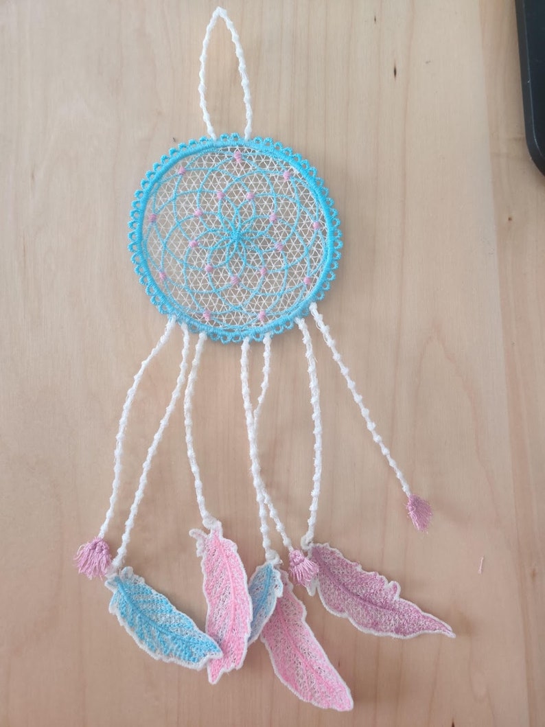 May include: A blue and white dreamcatcher with pink feathers hanging from it. The dreamcatcher is made of lace and has a beaded border. The feathers are made of fabric and have a delicate, lacy design.
