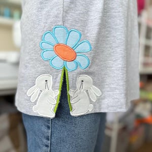 Può includere: Una maglietta grigia con un fiore blu e due coniglietti bianchi ricamati sull'orlo inferiore.