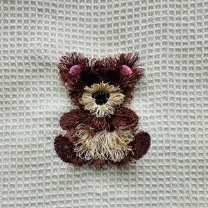 May include: A brown teddy bear with pink ears embroidered on a white waffle weave fabric.