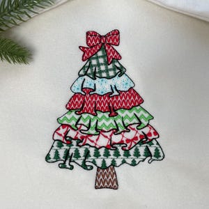 May include: A colorful Christmas tree applique with a red bow at the top. The tree features layers of green, red, and blue patterned fabric. The tree trunk is brown. The applique is on a white background.