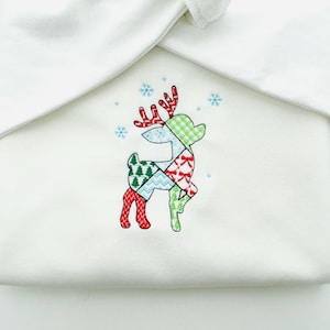 Könnte beinhalten: Ein weißes Stoffstück mit einem gestickten Patchwork-Rentier-Design. Das Rentier hat rote Geweihe, einen grün-weiß karierten Kopf und einen Körper, der aus verschiedenen gemusterten Flicken besteht. Blaue Schneeflocken sind um das Rentier herum gestickt.