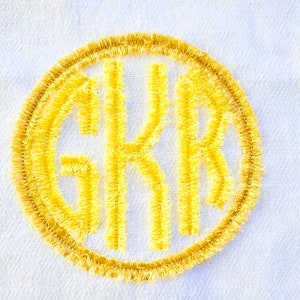 May include: A yellow embroidered monogram with the letters "GKR" in a circle on a white fabric background.