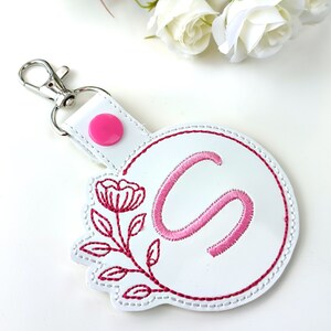 May include: White leather keychain with a pink embroidered floral design and the letter "S" in a circle. The keychain has a silver metal clasp.