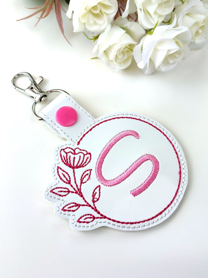 May include: White leather keychain with a pink embroidered floral design and the letter "S" in a circle. The keychain has a silver metal clasp.