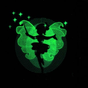 May include: A silhouette of a fairy with outstretched arms, wings, and a wand, surrounded by a green glow and stars. The fairy is centered within a circular design, all against a black background. The image has a whimsical, magical theme.