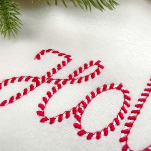 May include: A white fabric surface featuring red and white candy cane striped embroidery that spells out the word HOI in a cursive script font, with a small portion of a green pine branch visible at the top edge of the frame.