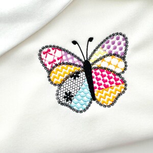 May include: Embroidered butterfly design with a patchwork pattern in various colors, including pink, yellow, purple, and blue, on a white fabric. The butterfly has a black body and antennae, and a black lace border.