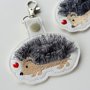 May include: White leather keychain with a gray and brown embroidered hedgehog with a red heart. The keychain has a silver metal ring.