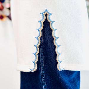 May include: A white garment featuring a side slit with a decorative scalloped edge. The embroidery along the opening consists of a light blue outer border and a tan inner border. The garment is layered over dark blue denim fabric.