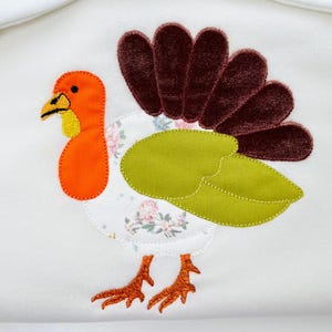 May include: A white fabric item featuring an appliqued turkey design. The turkey has an orange head and feet, a yellow beak and wattle, and a brown velvet tail. The body is white with a floral pattern and green leaves.