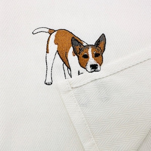 May include: White herringbone fabric featuring an embroidered dog. The dog is brown and white with black detailing. The dog is standing and looking forward. The fabric is folded over.