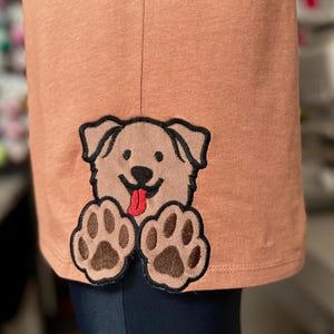 May include: A peach-coloured t-shirt with an embroidered brown dog peeking out from the bottom hem. The dog has a red tongue sticking out and brown paw prints.