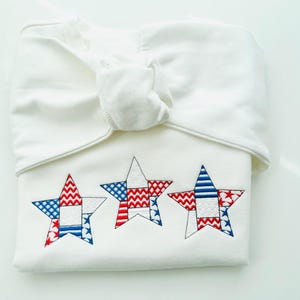 May include: A folded white baby outfit with three embroidered stars in red, white, and blue. The stars have a patchwork design with various patterns. The outfit is tied at the top with a knot.