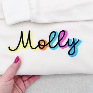 Könnte beinhalten: Cremefarbenes Sweatshirt mit dem Namen "Molly" in einer farbenfrohen, verspielten Schriftart gestickt. Die Buchstaben sind schwarz umrandet und mit gelben, orangefarbenen, rosa und blauen Akzenten versehen. Das Sweatshirt ist gefaltet, mit einem weißen Kragen sichtbar.