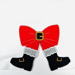 以下が含まれることがあります： 白い生地に、金色のバックルが付いた赤いリボンと、白いファーのトリムが付いた黒いブーツが刺繍されています。