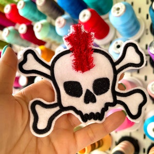 May include: A white felt skull and crossbones patch with a red felt mohawk. The patch is held in a hand against a background of colorful spools of thread.