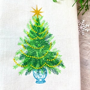 Op de afbeelding: Geborduurde kerstboom met een gouden ster bovenop, versierd met gele lichtjes en geplaatst in een blauw-witte vaas.