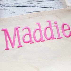 May include: A white canvas apron with the name "Maddie" embroidered in pink.