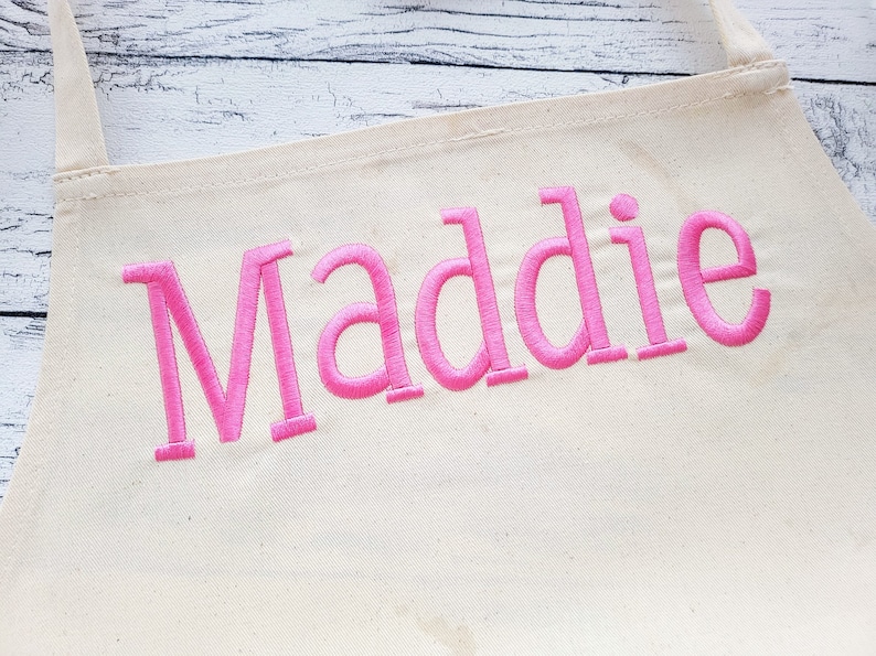 May include: A white canvas apron with the name "Maddie" embroidered in pink.