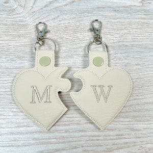 Puede incluir: Dos llaveros de color crema en forma de corazón con un diseño de pieza de rompecabezas. Los llaveros están unidos a llaveros de plata. El llavero de la izquierda tiene la letra "M" bordada, y el llavero de la derecha tiene la letra "W" bordada.