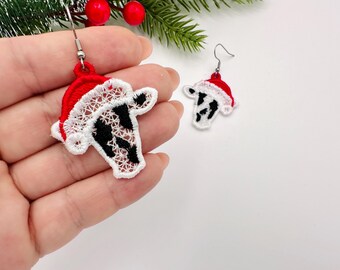 May include: A pair of festive Christmas earrings. Each earring features a red Santa hat with white trim, and a black and white cow face design. The earrings are made of lace and have silver-colored hooks.