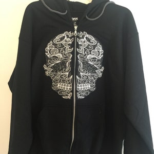 Pode incluir: Casaco de moletom preto com z&iacute;per e capuz, com um design de caveira de a&ccedil;&uacute;car bordada em branco na frente. O capuz &eacute; cinza e tem um cord&atilde;o.