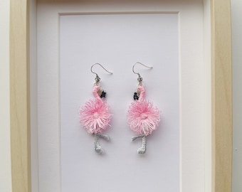 May include: A pair of pink flamingo earrings with silver hooks. The flamingos are made of pink and white thread and have gray legs.