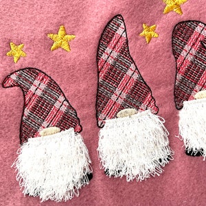 Puede incluir: Tres gnomos bordados con sombreros de cuadros rojos y negros y barbas blancas y peludas sobre un fondo rosa con estrellas amarillas.