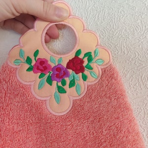 May include: A peach-coloured kitchen towel with a floral embroidery design. The embroidery features three pink, purple, and red roses with green leaves. The towel has a loop for hanging.