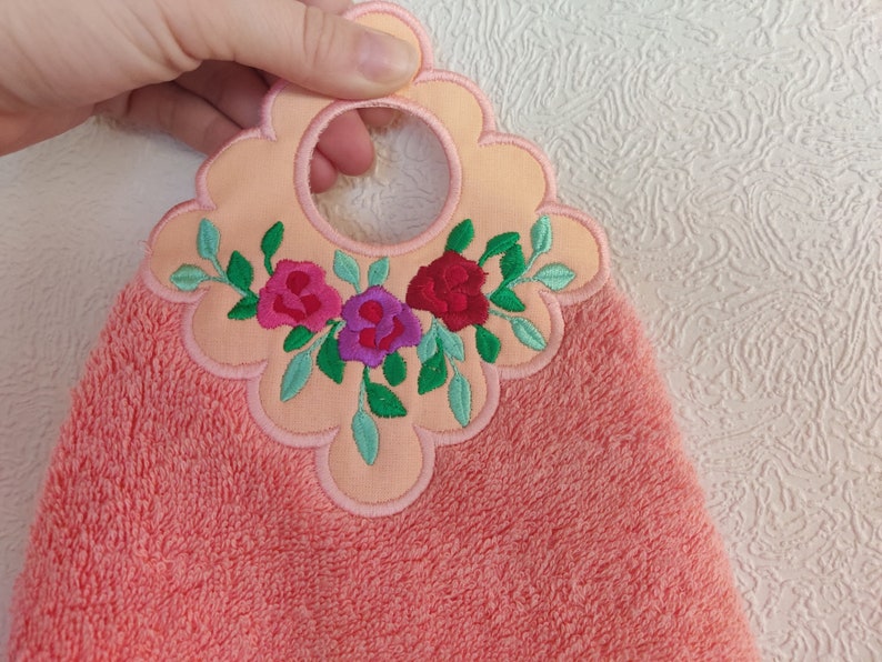 May include: A peach-coloured kitchen towel with a floral embroidery design. The embroidery features three pink, purple, and red roses with green leaves. The towel has a loop for hanging.