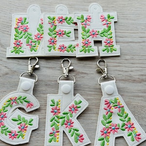 May include: Five white keychains with floral embroidery. Each keychain features a different letter of the alphabet, 'M', 'E', 'H', 'G', and 'K'. The letters are embroidered with pink, green, and blue flowers.
