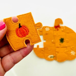 May include: A felt pumpkin puzzle with orange and yellow pieces. One piece is held up, showing an embroidered orange pumpkin with a black stem. The other pieces form a pumpkin shape with embroidered details.