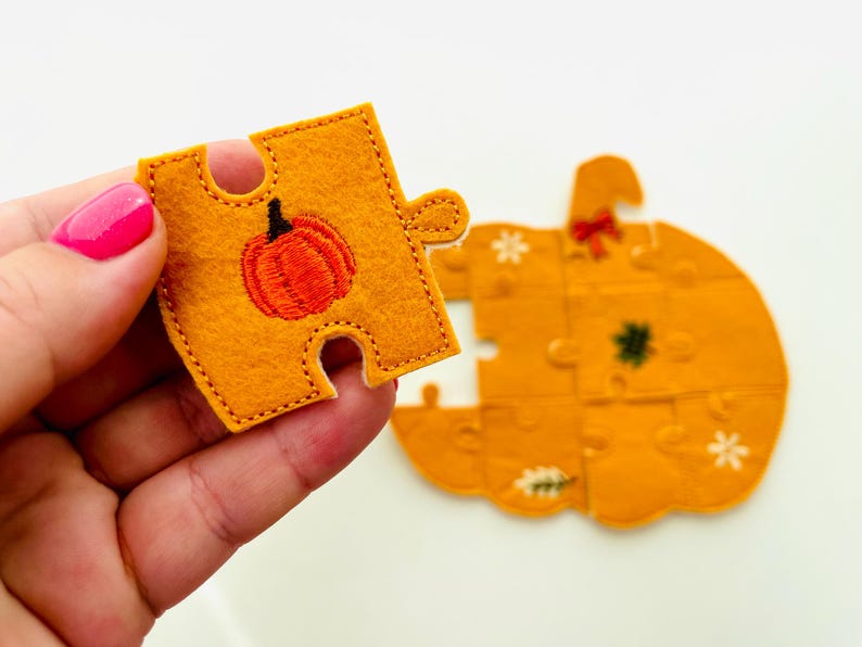 May include: A felt pumpkin puzzle with orange and yellow pieces. One piece is held up, showing an embroidered orange pumpkin with a black stem. The other pieces form a pumpkin shape with embroidered details.
