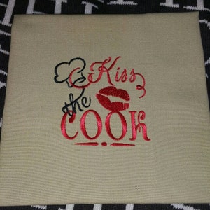 Może przedstawiać: Beżowy kwadratowy ręcznik kuchenny z haftowanym tekstem i grafiką. Tekst brzmi "Kiss the Cook" w czerwonym skrypcie, z czerwonym nadrukiem warg i czarną grafiką czapki kucharskiej.