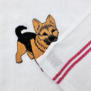 May include: White kitchen towel with an embroidered German Shepherd dog. The dog is depicted in shades of brown, black, and tan. The towel has a red striped border.