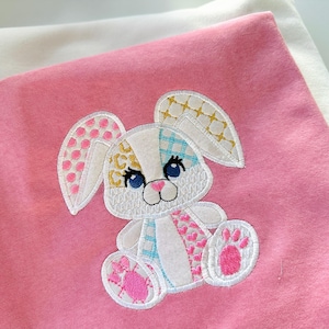 May include: A pink and white patchwork bunny applique with blue, yellow and pink accents. The bunny is sitting on a pink fabric background.
