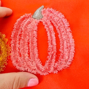 May include: Close-up of an orange fabric with embroidered pumpkin designs. One pumpkin is pink with a silver stem, and another is gold. The embroidery has a fringed texture, creating a 3D effect.