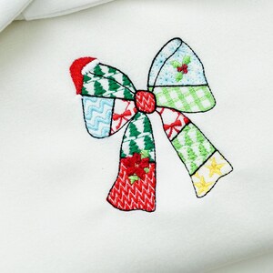 K&ouml;nnte beinhalten: Gestickte Weihnachtsschleife mit Patchwork-Design. Die Schleife hat rote, gr&uuml;ne, blaue und gelbe Abschnitte mit festlichen Mustern wie Weihnachtsb&auml;umen, Stechpalmen und Sternen. Die Schleife ist auf einem wei&szlig;en Hintergrund zentriert.