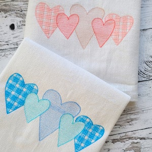 May include: Two white towels with embroidered rows of hearts. The top towel has pink hearts with a checkered pattern. The bottom towel has blue hearts with a checkered pattern.