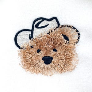 May include: Embroidered patch featuring a teddy bear wearing a cowboy hat. The bear has brown, textured fur and a black nose and eyes. The cowboy hat is black with a white brim. The patch is on a white background.