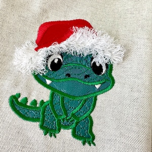 May include: Embroidered green alligator wearing a red Santa hat with white trim. The alligator has large black eyes and white teeth. The design is on a light beige fabric.